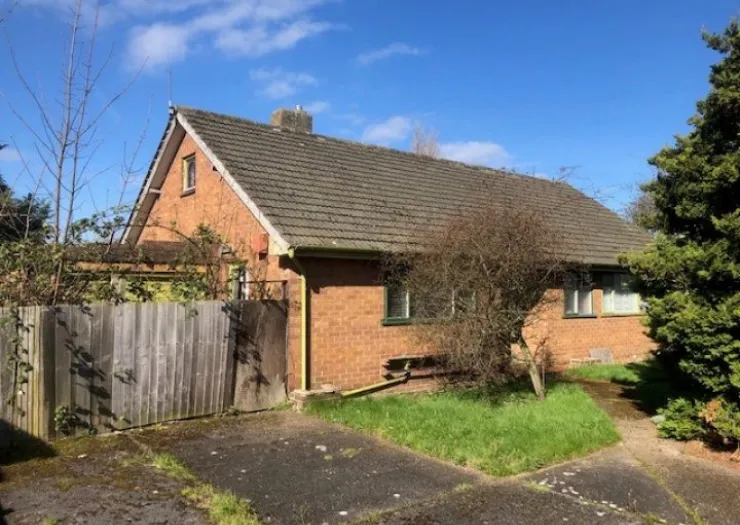 Brick bungalow exterior with tiled roof, fenced yard, and overgrown garden.
