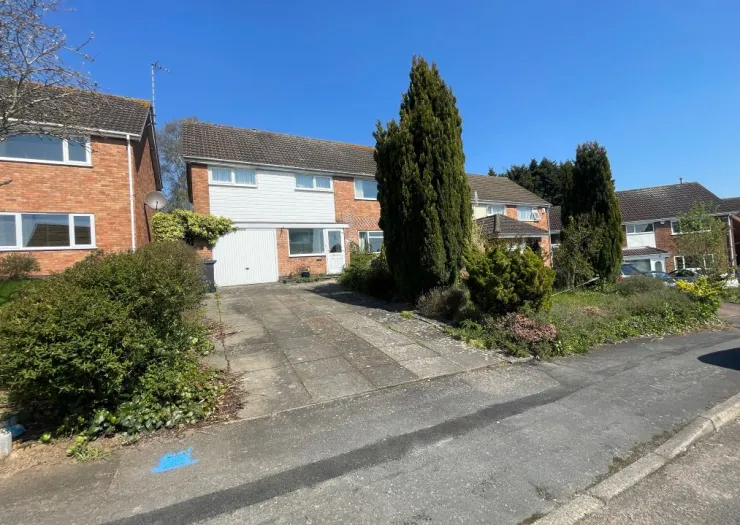 Suburban house exterior with driveway, front garden, and tall evergreen trees.