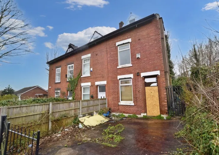 Red brick terraced house with boarded-up doorway and overgrown front yard.