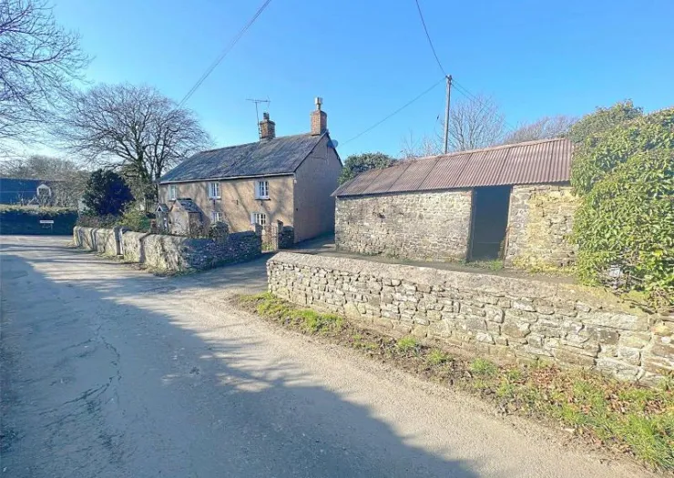 Charming rural cottage with attached stone barn and low stone wall along a quiet country lane.