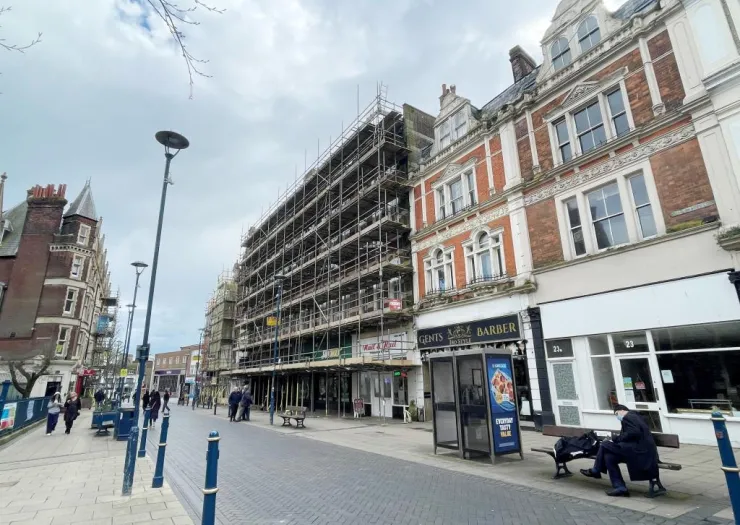 Scaffolding covers a building on a UK high street, pedestrians walk past shops and a man sits on a bench.