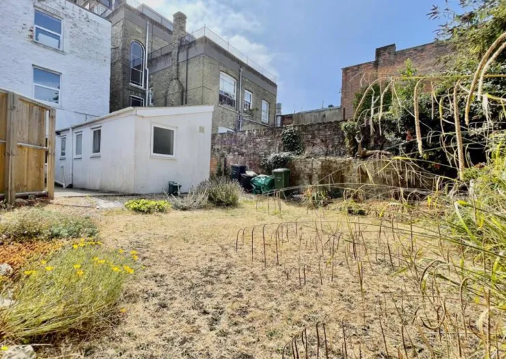 Overgrown garden with outbuilding, dry grass, and brick wall.