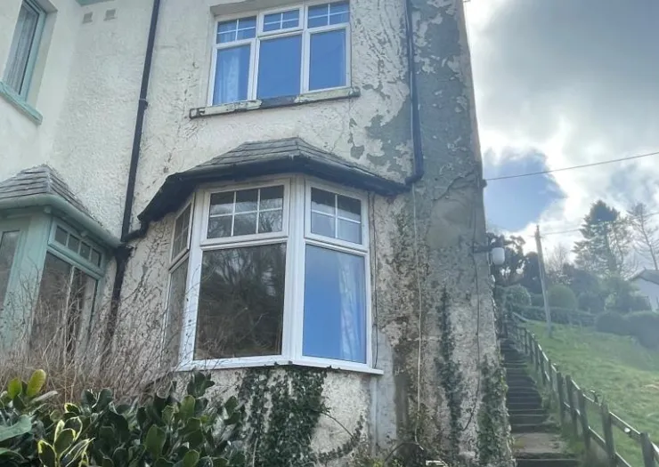 White bay window on house with peeling render, steps leading uphill to garden.