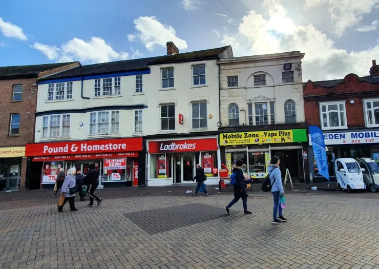 UK high street shops including Pound & Homestore, Ladbrokes, and Mobile Zone Vape Plus. Pedestrians walk on the paved street.
