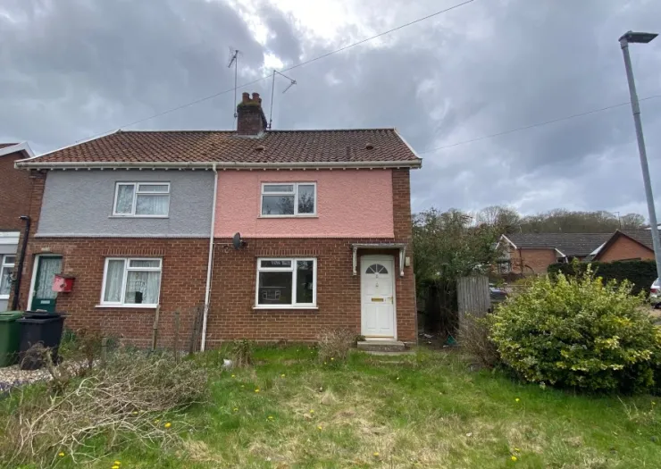 Pink and grey semi-detached house with front garden and overgrown bushes.