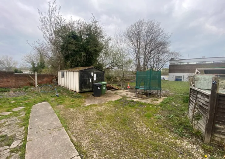 Overgrown garden with shed, trampoline, wheelie bins, and concrete path.