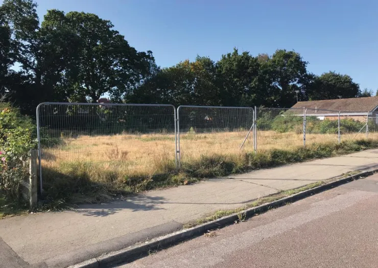 Empty fenced lot with overgrown grass, sidewalk, and trees.