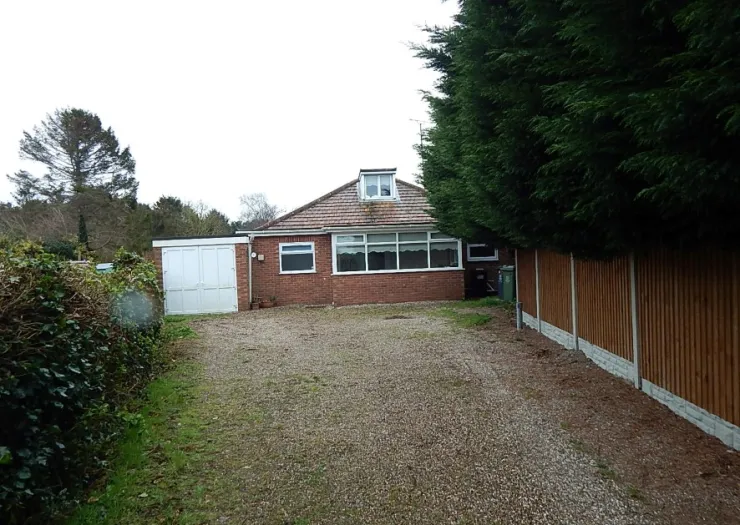 Red brick bungalow with attached garage and gravel driveway. Wooden fence borders the property.