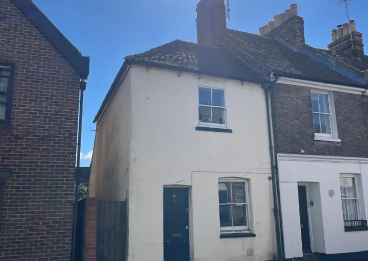 Small white terraced house exterior with black door and windows, between two brick buildings.