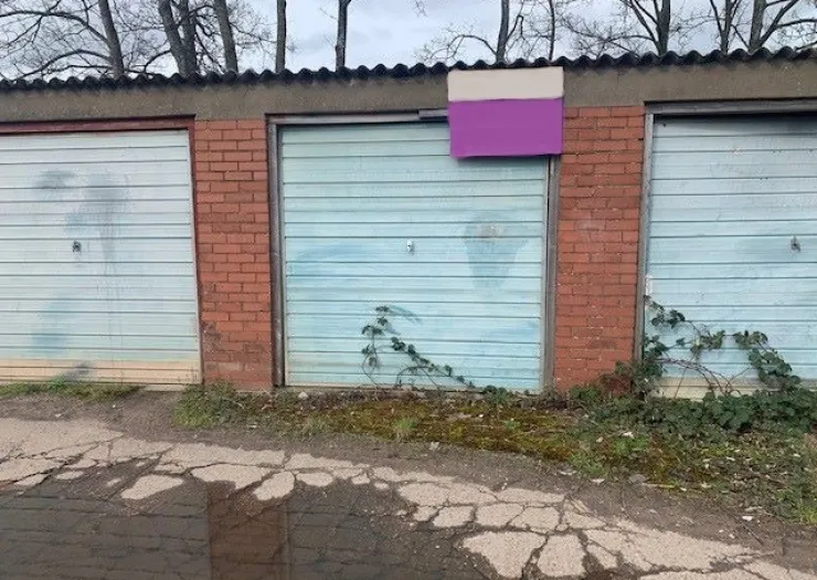 Row of light blue garage doors with brick dividers, some weeds, and a purple and cream sign on one door.