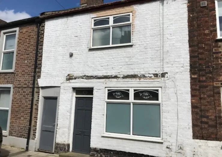 White painted brick terraced house with gray front door and frosted ground floor windows. Needs renovation.
