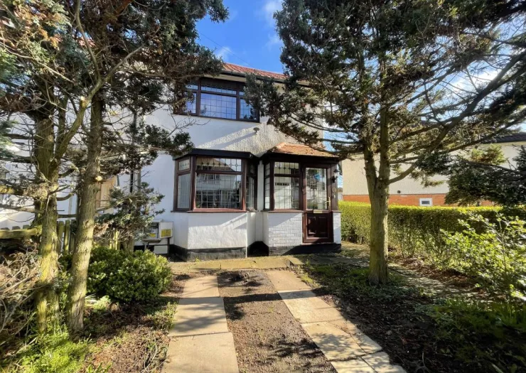 White detached house with dark brown window frames, paved driveway, and front garden with evergreen trees.