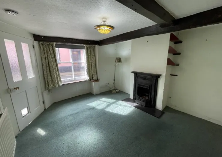 Empty living room with teal carpet, fireplace, exposed beams, and window with patterned curtains.