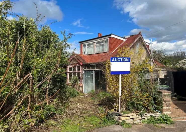 Overgrown detached house with "Auction" sign, needs renovation.