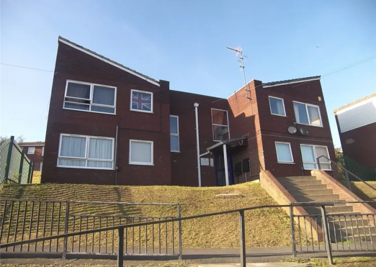 Red brick apartment building with sloped roofs, a Union Jack flag in the window, and a grassy front bank with railings.