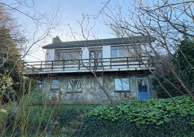 Two-story house with balcony, stone lower level, and surrounded by trees and greenery.