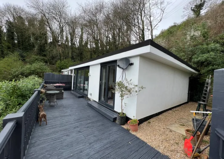 Modern white cabin with dark deck, patio furniture, and wooded hillside backdrop.