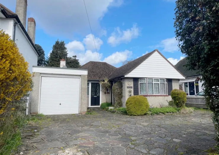 Suburban bungalow with attached garage, paved driveway, and front garden under a blue sky.