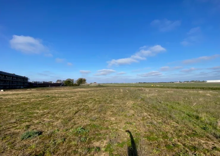 Empty field with sparse grass, a building under construction in the background, and a clear blue sky.