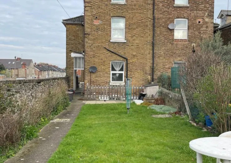 Back garden of brick house with lawn, shrubs, and wooden fence.