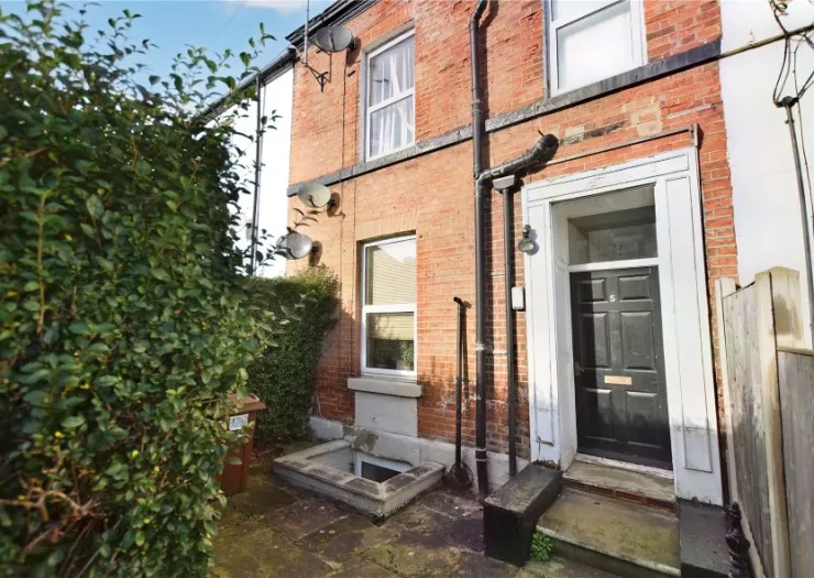 Brick house exterior with black front door, small paved yard, and green hedge.