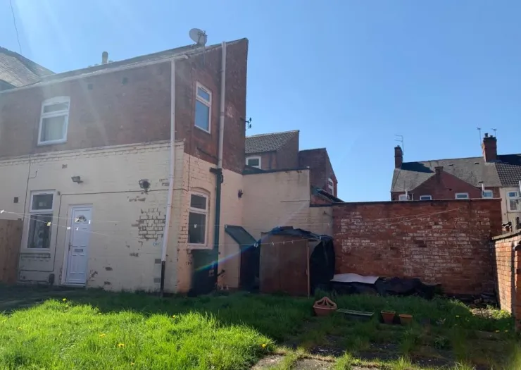 Backyard of a brick house with overgrown grass, a shed, and a brick wall.