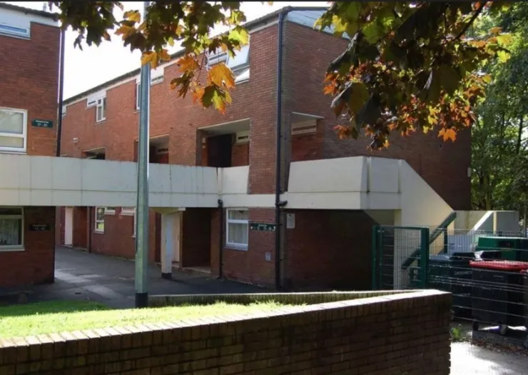 Red brick apartment building with a raised walkway, viewed from a low brick wall.
