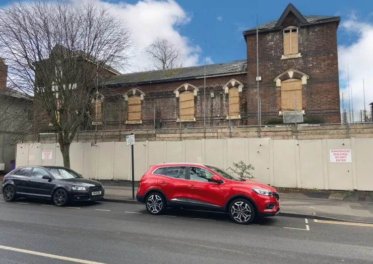 Boarded-up brick building behind a construction hoarding. "Dangerous Building. Do Not Enter" sign visible.