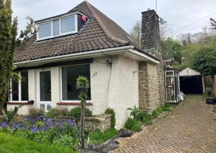 Suburban house with tiled roof, stone chimney, and brick driveway. Front garden with purple flowers. Needs renovation.