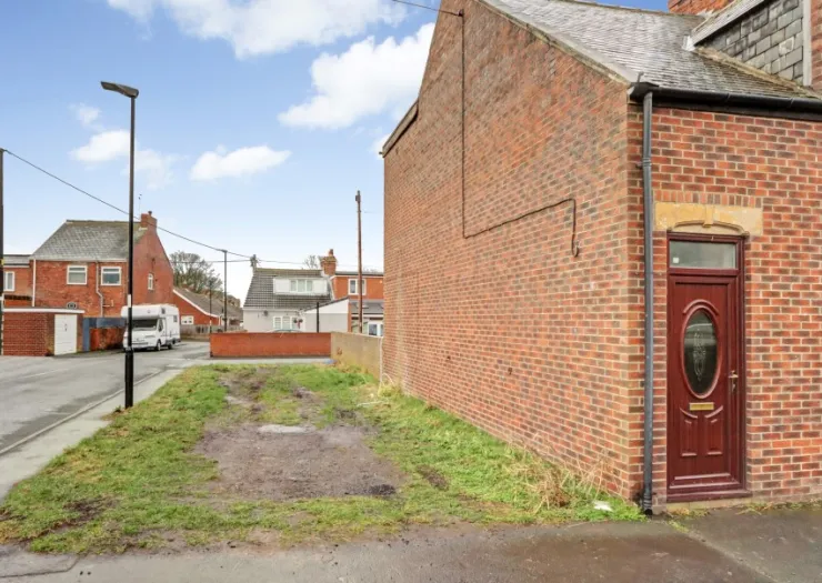 Brick house exterior with burgundy door beside a grassy patch and street view of other houses.