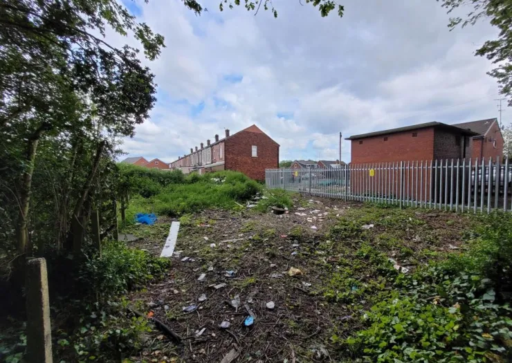 Overgrown vacant lot with litter, fenced area, and brick buildings in the background.