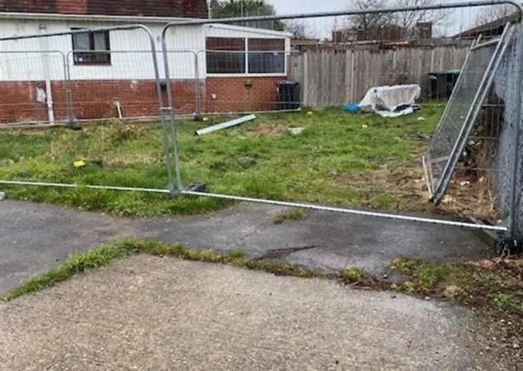 Overgrown grassy area behind metal fencing, with debris and a partially collapsed fence section.