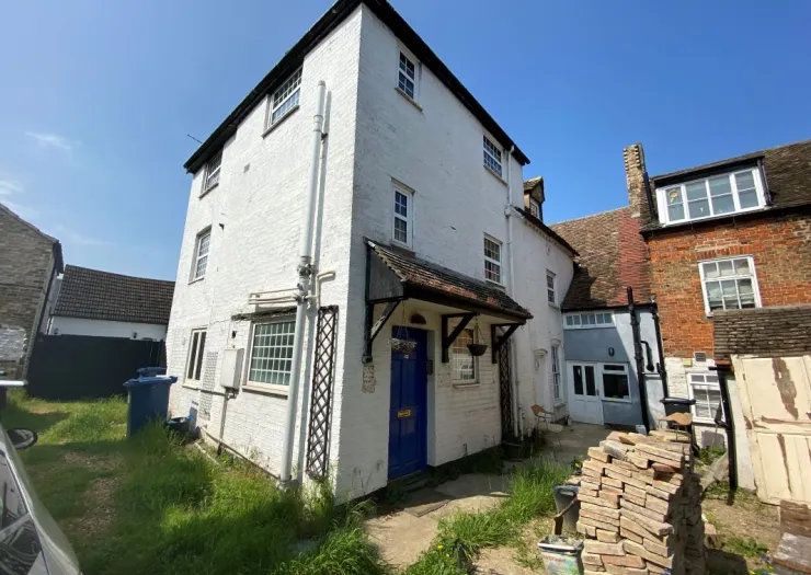 White, multi-story building with a blue door and small porch. Overgrown yard with a stack of bricks.