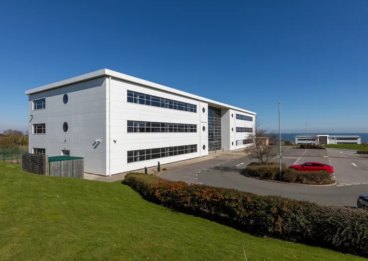 Modern white office building with parking lot, green lawn, and ocean view.