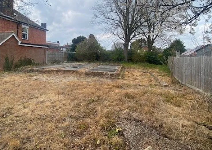 Overgrown lot with concrete foundation remnants, trees, and a partial view of a brick house.
