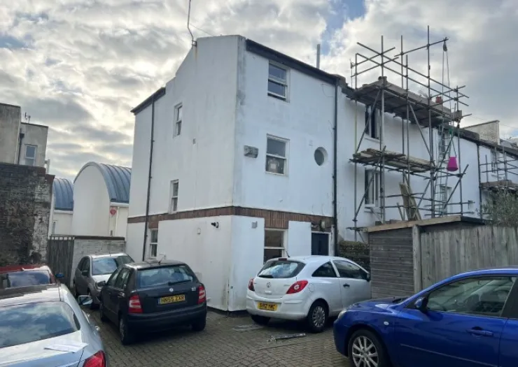 White building with scaffolding, small circular window, and parked cars in front.