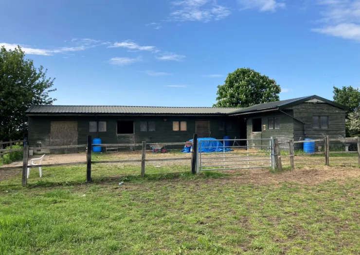 Green stable building with fenced paddock, blue tarp, and wheelbarrow.