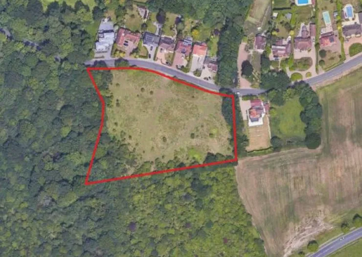 Aerial view of vacant land outlined in red, bordering a road and houses, surrounded by trees.