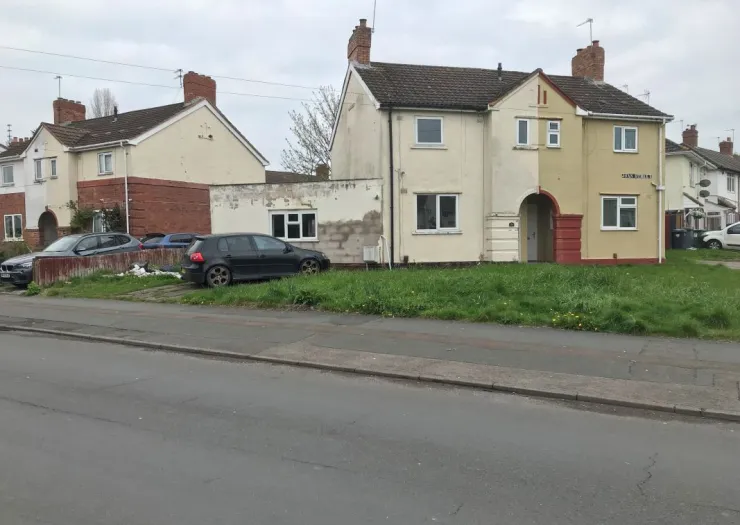 Semi-detached house with partially rendered exterior, small front lawn, and parked cars.