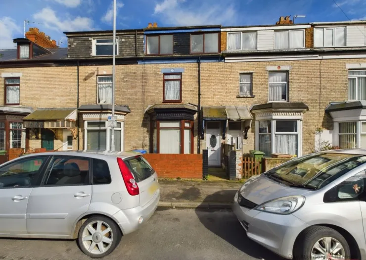 Exterior of a tan brick terraced house with bay window, parked cars on street.