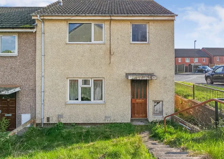 Exterior view of a two-story terraced house with a weathered facade, overgrown front garden, and steps leading to the brown f