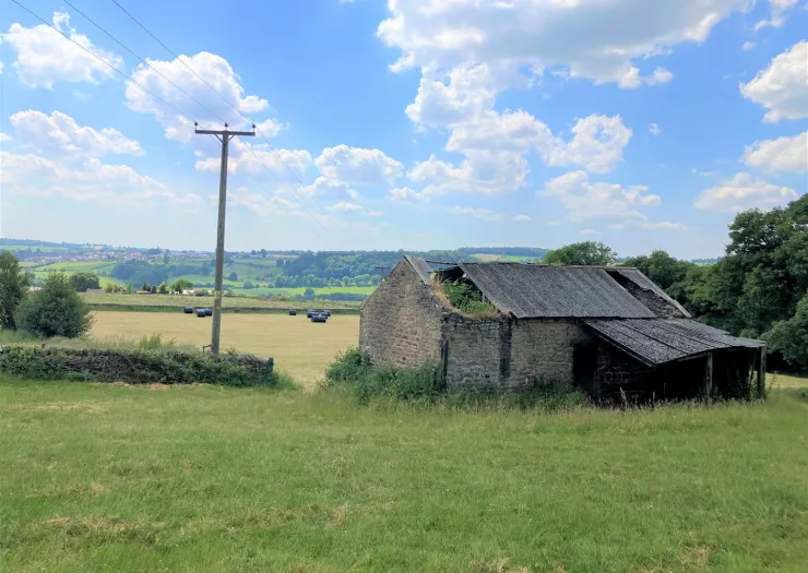 Dilapidated stone barn in a green field, with hay bales and rolling hills in the distance under a blue sky.