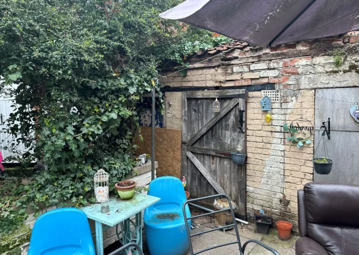 Small paved patio area with seating, a table, and overgrown shrubs next to an old brick outbuilding.