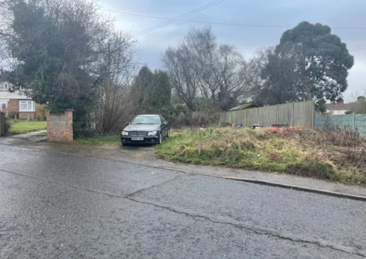 Overgrown plot of land beside a road with a parked car, trees, and fences.
