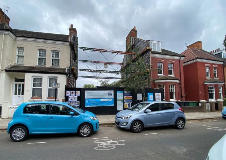 House under construction between two existing terraced houses, supported by scaffolding. Construction site signage visible.