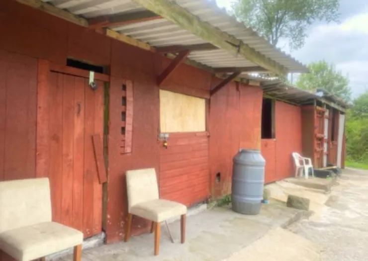 Red wooden outbuildings with boarded-up windows, a water barrel, and chairs outside.