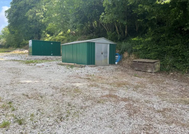 Two green metal storage sheds on gravel area near trees.