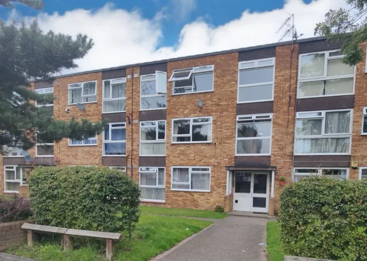 Brick apartment building exterior with white windows and shrubs under a blue sky.