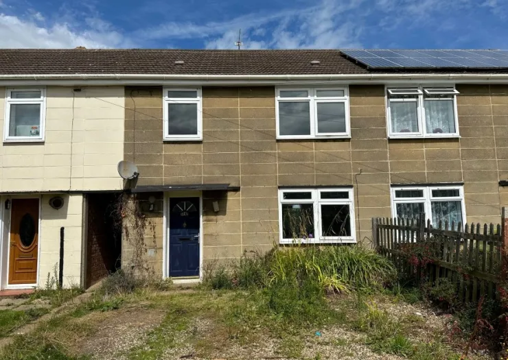 Attached house with solar panels, white windows, and a blue front door. Overgrown front garden.