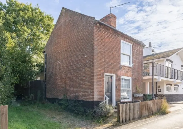 Charming red brick cottage with white trim, wooden fence, and neighboring building with balcony.